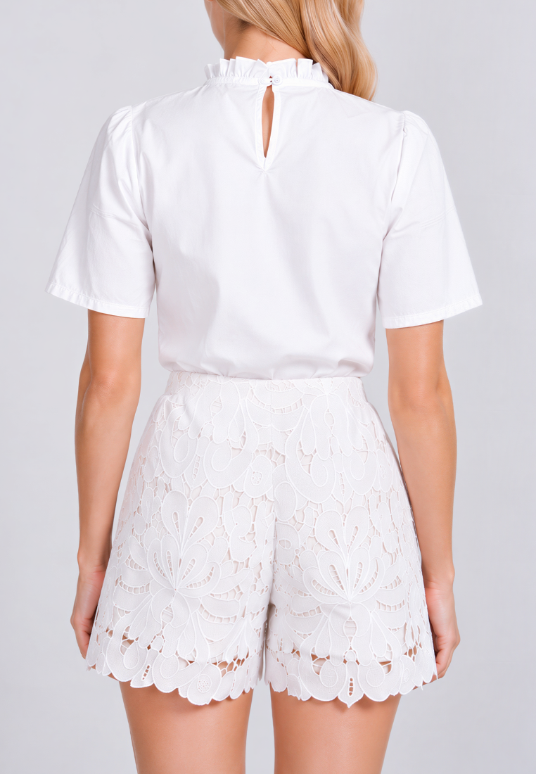 A woman shown from the back wearing a white short-sleeve blouse with a ruffled high neck and keyhole back detail, paired with white lace shorts, against a light gray studio background.