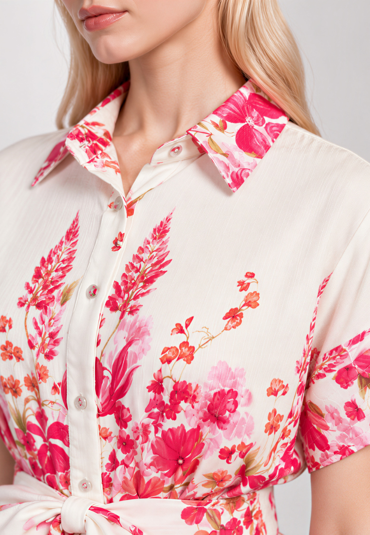 A close-up of a woman wearing a short-sleeve collared shirt dress with a vibrant pink and red floral print on a cream background.