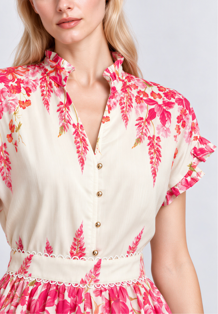 A close-up of a woman wearing a cream-colored short-sleeve button-front dress with a vibrant pink and red botanical leaf print, featuring a ruffled collar and sleeves