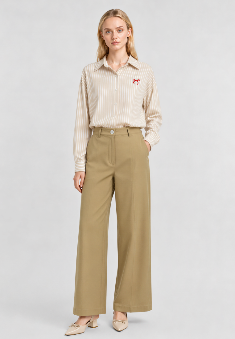 A woman stands against a light gray studio background wearing a beige and white vertically striped button-down shirt with a small red embroidered logo on the chest, paired with high-waisted khaki wide-leg trousers and beige pointed-toe heels.