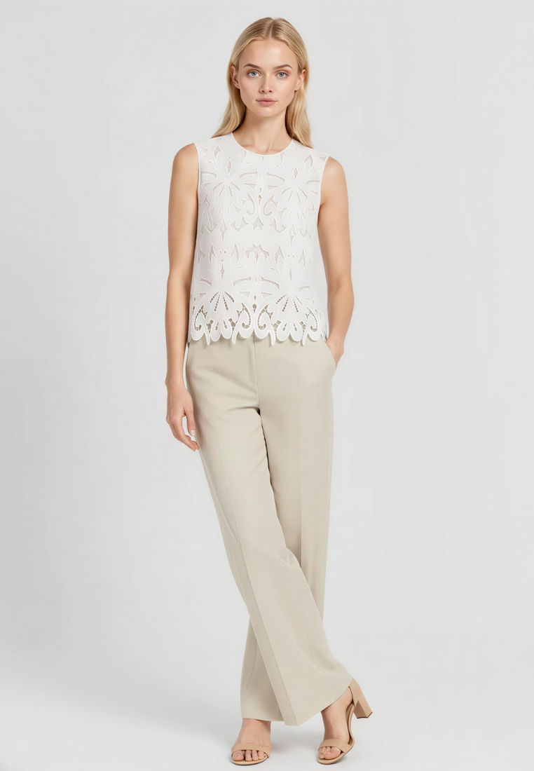 A woman modeling a sleeveless white top with intricate floral eyelet embroidery and a scalloped hem, paired with light beige wide-leg trousers and nude heeled sandals, against a light gray studio background.