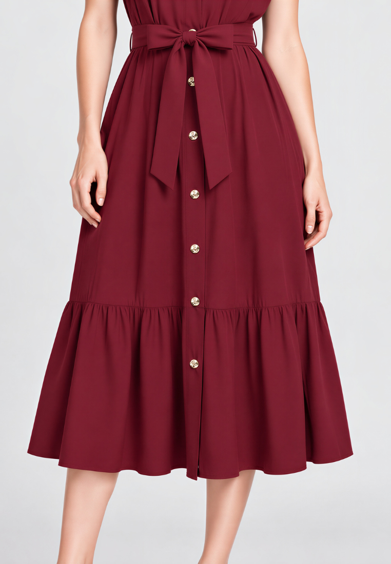 A woman wearing a sleeveless burgundy midi dress with a tie waist, front button closure, and a ruffled hem, shown against a light gray studio background.