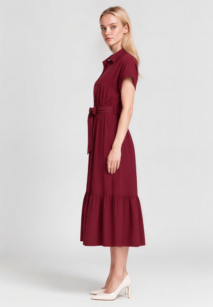A woman modeling a short-sleeve midi dress in deep burgundy with a matching waist tie, paired with white pointed-toe heels, against a light gray studio background.