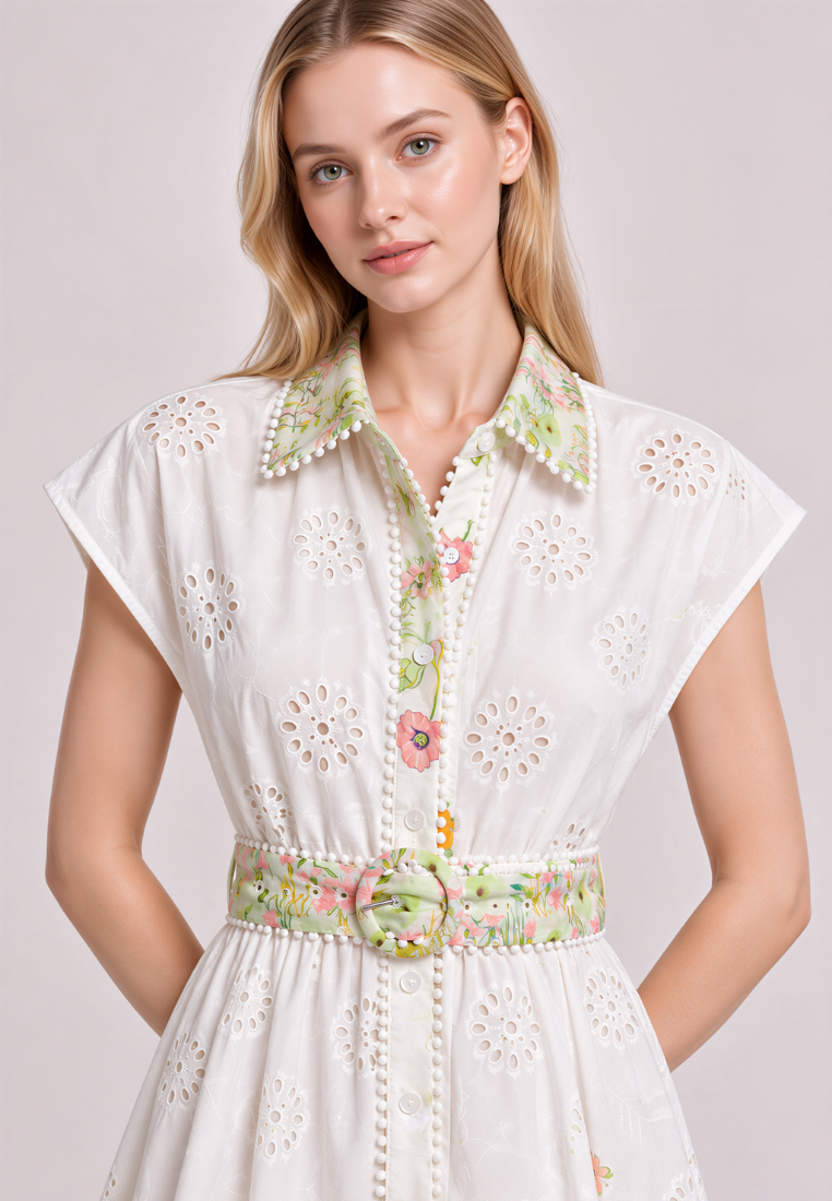 A woman wearing a white eyelet dress with a floral print collar, button placket, and matching floral belt, photographed against a light gray studio background.