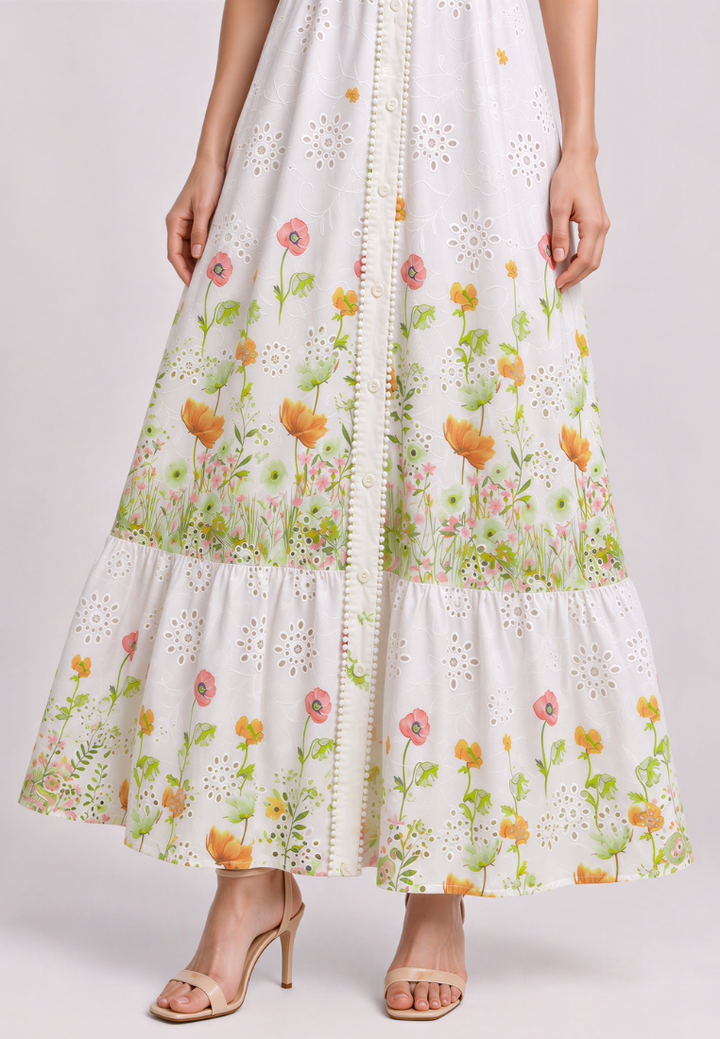 A white tiered maxi dress with a colorful floral print and embroidered eyelet details, shown on a model wearing beige high-heeled sandals against a light gray studio background.