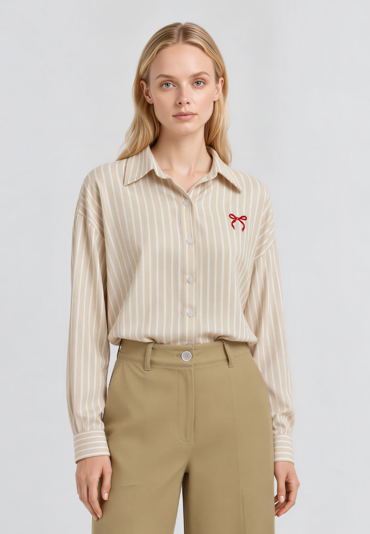A woman modeling a beige and white vertically striped button-down shirt with a small red bow embroidery on the chest, paired with khaki trousers, against a light gray studio background.