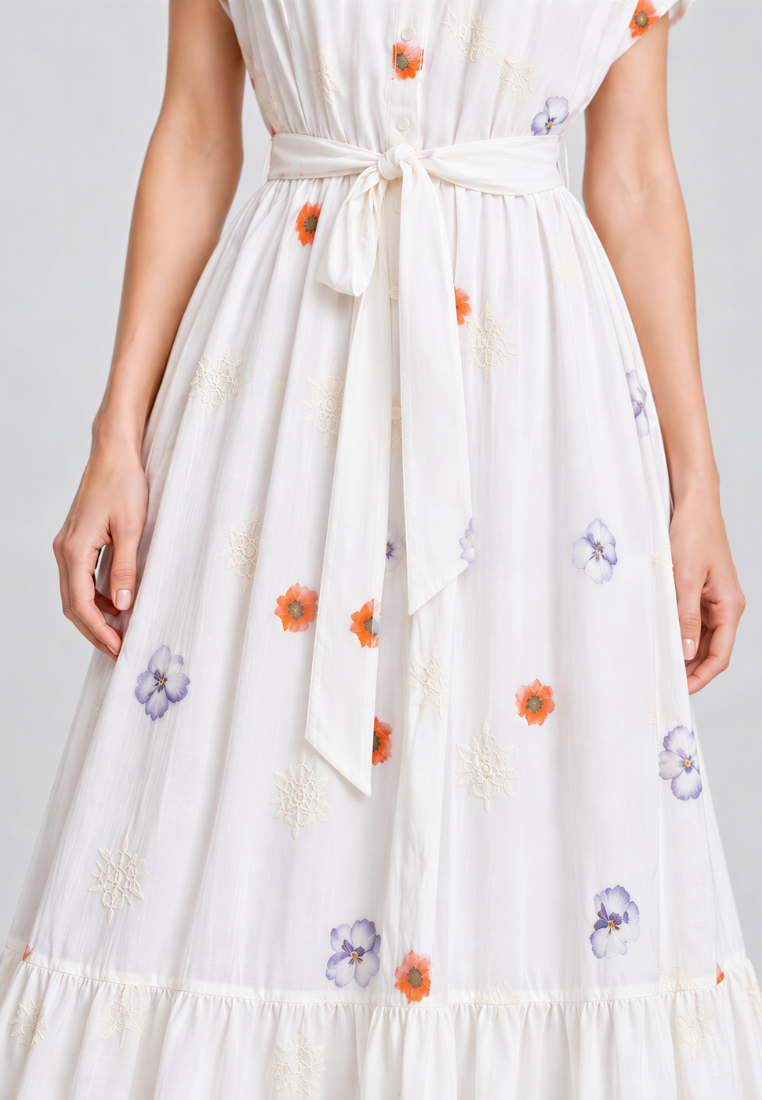 A close-up of a white tiered maxi dress with a matching waist tie belt, featuring embroidered orange and purple flowers and white starburst motifs.