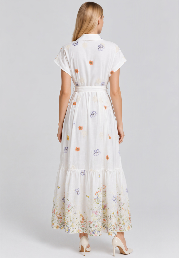 A woman viewed from the back wearing a white short-sleeve maxi dress with a matching waist tie and a multicolor floral print, paired with beige high heels against a light gray studio background.