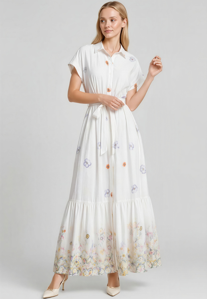 A woman modeling a white short-sleeve maxi shirt dress with a tiered skirt and delicate pastel floral embroidery, set against a solid light gray studio background.
