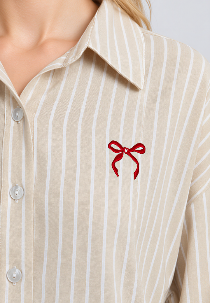 Close-up of a beige and white vertically striped button-down shirt worn by a model, featuring a small red embroidered bow on the left chest.