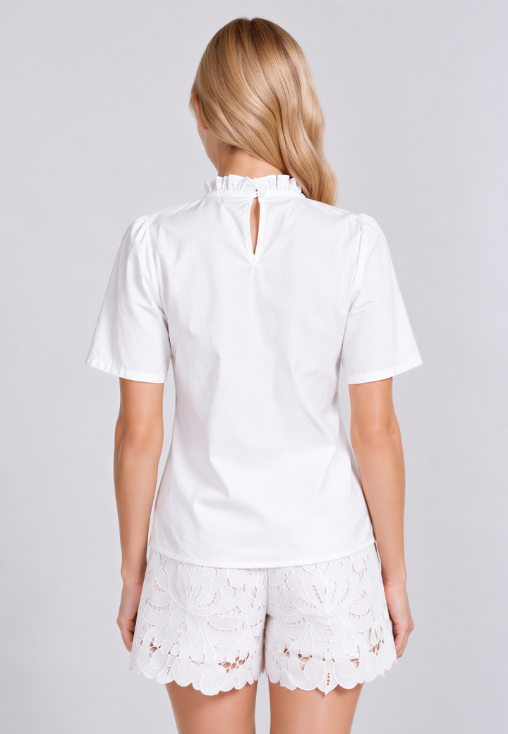 A woman shown from the back wearing a white short-sleeve blouse with a ruffled high neck and keyhole back detail, paired with white lace shorts, against a light gray studio background.
