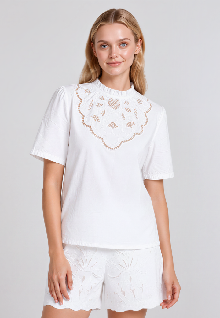 A woman modeling a white short-sleeve eyelet top with a scalloped floral neckline, paired with matching white eyelet shorts, against a light gray studio background.