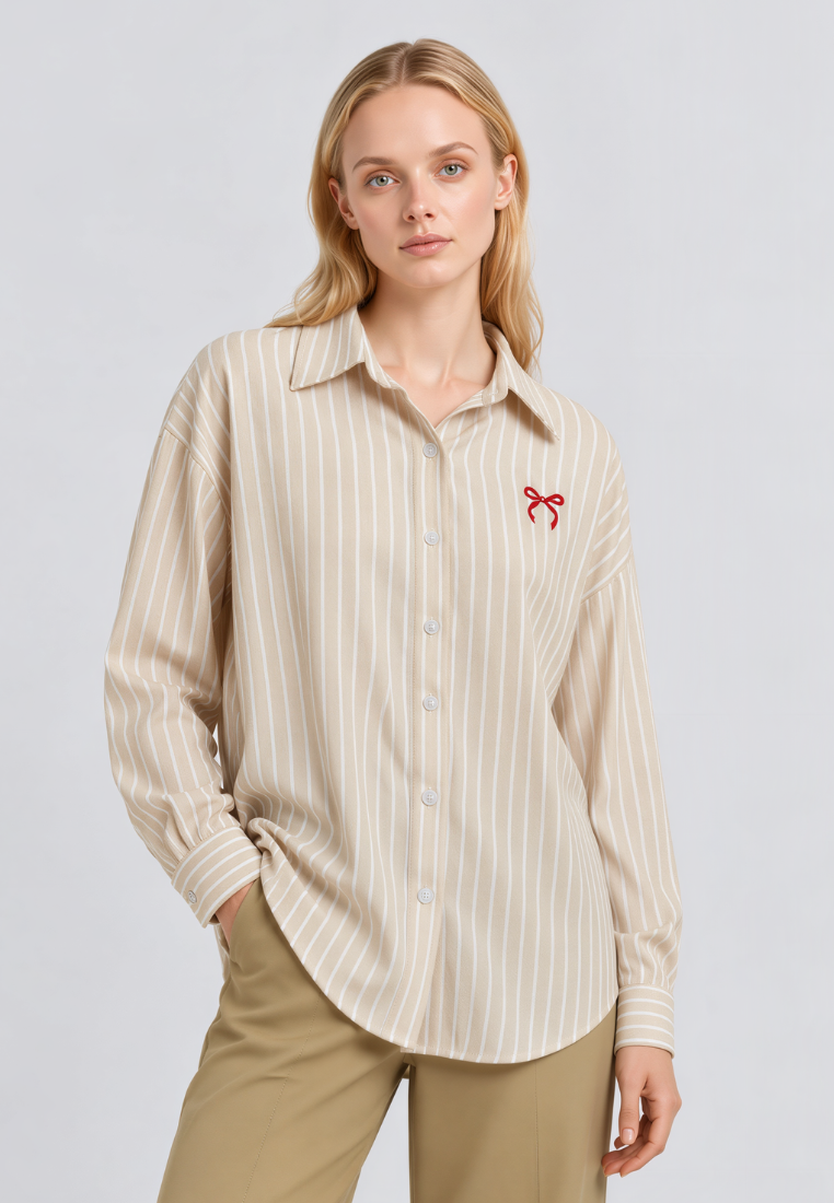 A woman modeling a beige and white vertically striped button-down shirt with a small red bow embroidery on the left chest, paired with khaki pants, against a light gray studio background.