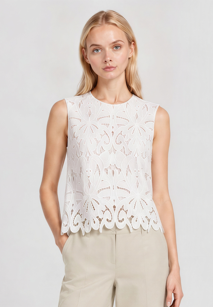 A blonde model wearing a sleeveless white eyelet lace top with a scalloped hem and light beige trousers, photographed against a light gray studio background.