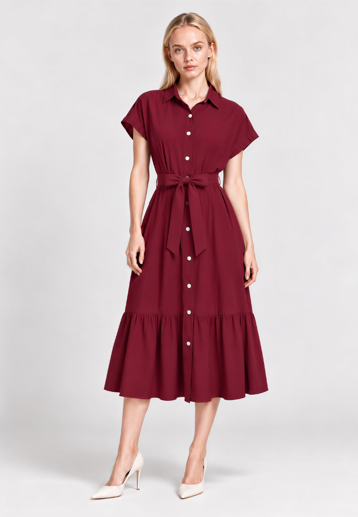 A woman modeling a short-sleeve midi dress in deep burgundy with a button-down front, a matching waist tie, and a ruffled hem, paired with cream high heels against a light gray studio background.